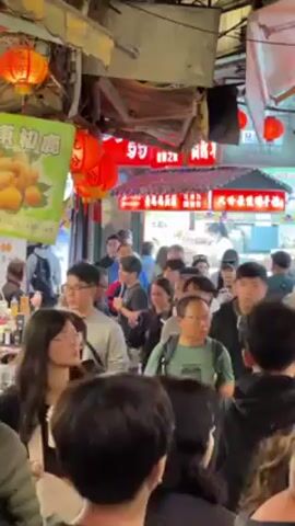 The video shows a crowded outdoor market scene. A diverse group of people, mostly appearing to be of Asian descent, are walking through a narrow corridor. Many people are wearing glasses and masks. There are red lanterns and commercial signs hanging overhead, indicating a lively, possibly festive atmosphere. The camera is moving with the crowd, capturing the density and bustle of the marketplace.