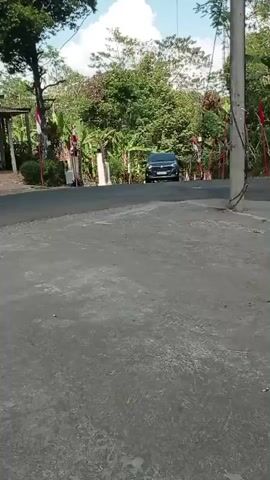 A black car drives down a paved road lined with trees and houses. The car is seen from the side, and it is moving away from the camera. There are flags on poles on either side of the road. The sky is blue with some clouds. The video is shaky, suggesting it was recorded by someone walking or in a moving vehicle.