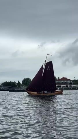 A wooden sailboat with maroon sails glides across choppy water under a cloudy sky. The sails display the number "8" and the letters "GH." Several people are visible on board the boat. In the background, a shoreline with trees and buildings can be seen.