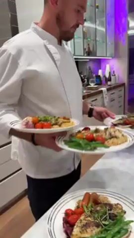 A chef wearing a white chef's coat and black pants carries a stack of plates of food from the kitchen to a group of people seated at a table in the dining area of an upscale residence. The plates contain a piece of grilled fish, rice, green beans, cherry tomatoes, and greens. The kitchen has white cabinets and a marble countertop where several plates are arranged. The dining area is lit by indirect lighting, with a modern chandelier. The dining area is open to a living room with a large television. The video captures the chef walking towards the seated guests with the plated meals.