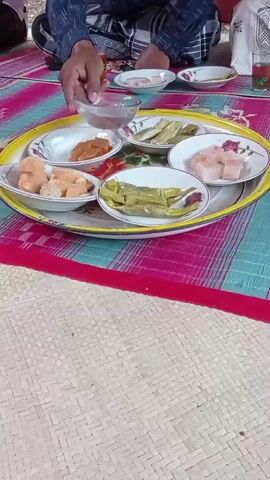 The video shows a close-up of a tray with several small plates of food. The tray is yellow and has a floral design around the edge. The plates contain what appears to be small pieces of fried food, some green items that might be vegetables or fruit, and some cubed white food. A hand is visible reaching into one of the plates with a spoon or fork. In the background, a person is wearing a patterned shirt and a sarong. The tray is placed on a mat with a checkered pattern.