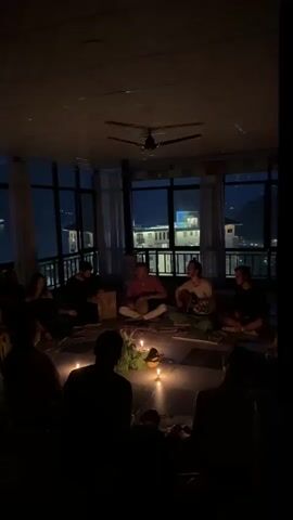 A group of people are gathered in a dimly lit room, sitting on the floor in a circle around lit candles and foliage. Five people are positioned closer to the camera than the others, and these five are playing instruments. One is playing a stringed instrument, possibly a guitar. One is playing what appears to be a drum held in their lap. The windows behind them show a night scene of a building and city lights. A ceiling fan is visible in the upper center of the frame.