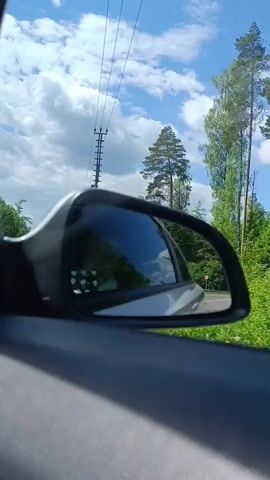 The video shows a car's side mirror while driving on a road bordered by green trees. The sky is blue with white clouds. The mirror reflects the car behind and the passing landscape. There's a utility pole visible next to a tall pine tree.  Music is playing in the background. The shot is taken from inside the car.