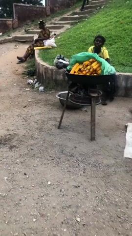 The video shows an outdoor scene, possibly a street or market. In the foreground, there is a dirt path with scattered litter. A woman is seated on a low wall to the left, wearing a patterned dress and a headscarf. Next to her, on the ground, are some bags. In the center of the frame, a charcoal grill with a large bowl of corn on the cob is set up. Another woman, wearing a yellow top, is seated to the right of the grill. Further back, there are concrete steps leading up a grassy slope, with a few people walking up them. A man in a blue patterned outfit is walking away from the camera on the dirt path, carrying two bags. The overall environment appears to be somewhat unkempt, with visible debris and vegetation.