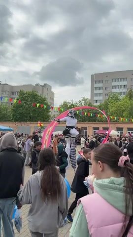 A stilt-walker dressed in black and white jester costume performs in a town square packed with people. The stilt-walker manipulates long, pink fabric pieces in the air. The sky is overcast. Behind the crowd, several apartment buildings are visible. Colorful pennants are hung across the square. People stroll past, some holding balloons or enjoying ice cream. The ground is paved with light-colored bricks.
