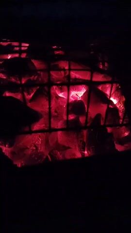The video shows a close-up, low-angle view of glowing red charcoal briquettes behind a black metal grill grate. The charcoal is intensely hot and emits a warm, orange-red light that illuminates the scene. Some of the briquettes appear to be actively burning, with small flames visible. The background is dark and out of focus, suggesting the footage was taken at night or in a dimly lit environment. The camera is relatively still, with slight movements that might indicate hand-holding. The overall impression is one of heat, fire, and the raw energy of combustion.