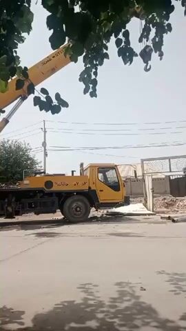 The video shows a yellow truck with a crane extended overhead. The truck is parked on a paved surface, possibly a street or construction site. Trees with green leaves overhang the scene from the top left. The sky is bright and clear. In the background, there is a white gate and some construction materials. The crane appears to be in operation, with its hook visible.