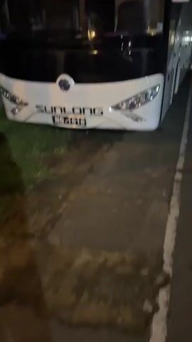The video is an extreme close-up shot of the front and side of a white bus, identified by the logo "SUNLONG" and the license plate "NG-1616" on the front. The bus appears to be parked on the side of a road at night, with streetlights and other vehicles visible in the background. The camera pans slowly along the side of the bus, revealing its wheels and the road surface. The overall impression is a still, observational shot of the bus in its environment.