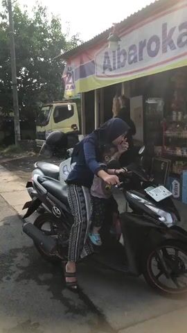 A woman wearing a blue hijab and a patterned outfit is sitting on a black scooter with a young child in front of her. The woman is holding onto the handlebars and the child is also touching the handlebars. They are parked on a paved surface next to a building with a sign that says "Albaroka." A yellow truck is visible in the background. The scene appears to be outdoors, possibly in a street or parking lot, with trees and sunlight. The scooter is positioned at an angle, facing slightly to the right of the frame. The woman is looking towards the child, and the child is looking forward.