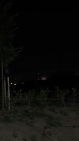 The video shows a nighttime scene with a dark, overcast sky. In the distance, a series of fireworks are set off, illuminating the sky with bursts of white, yellow, and pink light. The foreground is a dark, uncultivated field with a few scattered trees and saplings. The atmosphere appears calm and festive, likely a celebration.