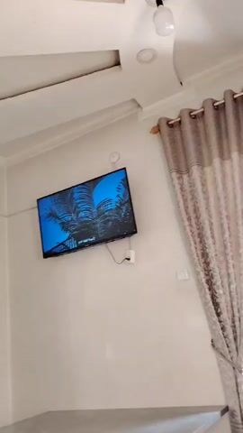 The video shows a wall-mounted television displaying text on a blue background. To the right of the television, a curtain hangs, covering a portion of the wall. The ceiling is visible at the top of the frame, with light fixtures. The overall scene appears to be an indoor setting, possibly a living room or a common area.