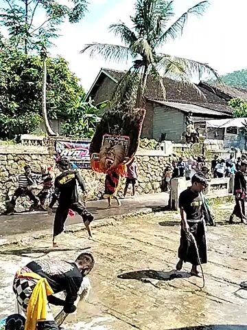 The video shows a traditional Indonesian performance taking place outdoors. A person wearing a large, elaborate mask and costume is dancing barefoot in the center of a paved area. The costume features a large headpiece adorned with feathers and intricate patterns, and the dancer is wearing a red and gold garment. Several other individuals are standing around the dancer, some dressed in black shirts and pants, and others in more casual attire. A man in the foreground is crouched down, possibly adjusting equipment or performing a task related to the performance. Behind the performers and spectators is a stone wall. A banner with colorful lettering is hung on the wall. Beyond the wall, there are houses, trees, and a palm tree visible against a partially cloudy sky. The ground is wet, suggesting it may have rained recently.
