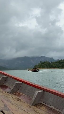 The video shows a view from a wooden boat moving on a body of water. In the distance, another longboat carrying people is visible. The background features lush green mountains shrouded in clouds, suggesting a tropical or mountainous region. The sky is overcast with heavy gray clouds. The water is a greenish-blue color and appears slightly choppy. The wooden boat in the foreground has red trim and visible planks. The overall atmosphere is moody and atmospheric due to the cloudy weather.