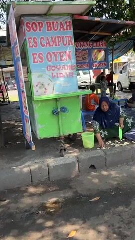 A woman wearing a hijab and a patterned dress is kneeling on the ground, cleaning fruit with a green bowl and a brush. She is in front of a food stall with a sign that reads "ES BUAH GOYANG LAMA". There is a bucket next to her and some fruit on the ground. In the background, there is a man sitting on the ground. The scene appears to be outdoors, possibly on a street or sidewalk.