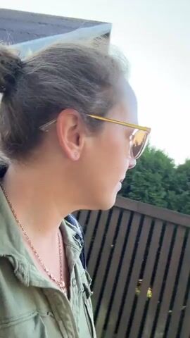 A woman is walking outdoors, her face is in close up. She is wearing sunglasses and a green shirt. She is also wearing a necklace. In the background, there is a fence and a house. The sun is shining and casting shadows on the ground. The woman is looking at the camera and smiling.
