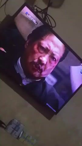 The video shows a television screen displaying a close-up of an Asian man's face. The man appears to be in distress, with a bruised and bloody cheek. He is wearing a dark suit and a white shirt. The lighting on the screen is dim and casts a purple hue on his face. In the foreground, out of focus, is a floor with some items on it, including what appears to be a box of tissues or wipes. The television is mounted on a wall, and some cables are visible behind it. The scene appears to be an indoor setting, possibly a living room. The video is of low quality and slightly blurry.