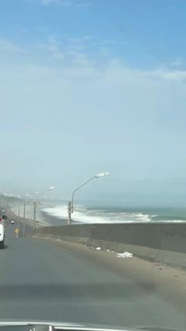 The video shows a view from inside a car driving along a coastal highway. To the right of the road is a concrete barrier, beyond which lies a beach with white waves crashing onto the dark sand. A row of tall, silver lampposts lines the road next to the beach. In the distance, the coastline extends into a misty horizon with faint outlines of buildings. The sky is light blue, with scattered clouds. Another car is visible in front and a motorcycle can be seen driving in the distance on the left side of the road. The quality of the video is clear, and the scene appears to be taken during the day.