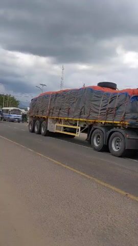 The video shows a traffic scene on an asphalt road running through an area that appears to be a town or settlement near a mountain range. The sky is overcast with grey clouds.

Initially, the view focuses on a large truck with a covered load moving from right to left. As the camera shifts, other vehicles come into view, including a yellow truck, a minibus, and a blue auto rickshaw. People can be seen walking on the sides of the road, near what seem to be small shops or stalls.

The camera angle changes slightly, revealing more of the road and the surrounding area. A bird flies across the screen. A motorcycle rider passes by, heading in the same direction as the trucks. The mountain range is visible in the background, adding context to the location.