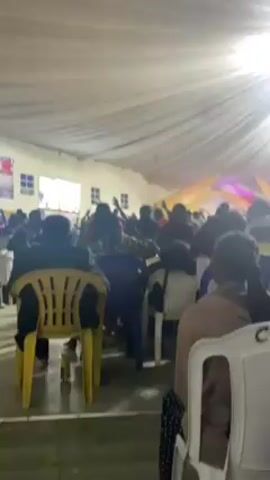 A low-angle shot inside a large tent shows a crowd of people with their hands raised in the air, presumably at a religious gathering or concert. The tent has a white fabric ceiling with folds. Stage lights illuminate a backdrop with gold and pink elements at the far end of the tent. People are sitting on plastic chairs in the foreground. A few windows are visible on the sides of the tent. The overall atmosphere suggests a lively, spiritual event.