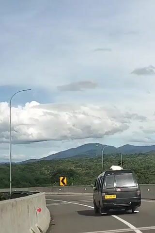 A dark colored minivan is driving on a highway. The minivan has a yellow license plate and a white object on its roof. The highway has concrete barriers on the left side and is bordered by green trees and hills in the background. There are streetlights and road signs indicating a curve. The sky is cloudy with some patches of blue.