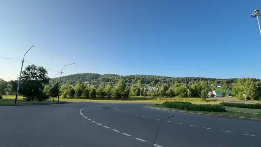 The video shows a road with a blue sky overhead and a grassy area with trees and a hill in the background. On the left side of the road is a tall brick apartment building. Further down the road, there are several trees and some buildings with colorful flags. A motorcycle is visible on the right side of the road. The road has white dashed lines indicating lanes. The overall scene suggests a rural or suburban environment on a clear day.