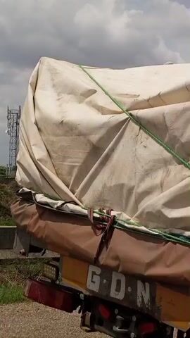 The video shows the back of a covered truck.  The cover is dirty and tied down with a green rope. The camera moves to the side and shows a white pick-up truck on the side of a road, with grass and bushes around it. There is a red building in the background.