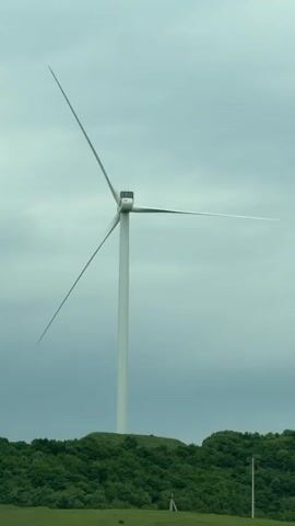 The video shows a white wind turbine standing on a small hill with green vegetation. The sky is overcast and grey. The camera appears to be moving, possibly from a vehicle on a road. In the foreground, there is a green field and a structure with multiple connected, white roofs. Loud music is playing in the background.
