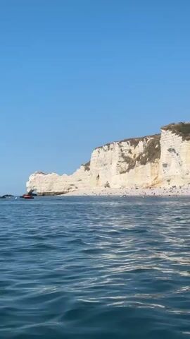 The video pans across a wide, bright blue sky. Below, a dramatic white cliff face rises from the ocean. The cliff is dotted with vegetation and has several caves or openings. At the base of the cliff, a sandy beach is visible, crowded with people sunbathing and walking. The ocean in the foreground is a deep blue with gentle waves. Further along the coast, the cliff continues, and on top of it, a small church with a steeple can be seen. A few buildings, including what appears to be a hotel with balconies, are situated on the hillside overlooking the beach. A red inflatable boat is visible near the cliff face on the left side of the frame. The overall scene depicts a sunny day at a popular beach resort.