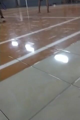 The video shows a badminton court with several people playing. The court has a wooden floor with white lines marking the boundaries. There are lights on the ceiling that illuminate the court. In the foreground, two people are playing badminton. One person is on the left side of the court, wearing a blue shirt and black shorts, and the other person is on the right side, wearing a white shirt and black shorts. There are other people in the background, some of whom are also playing badminton, while others appear to be waiting their turn. The game seems to be in progress, with players actively hitting the shuttlecock. The video is taken from a slightly elevated angle, looking down at the court. The lighting is bright, and the court appears to be well-maintained. There are also some spectators or other players seated on the side.