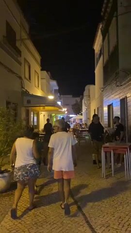 The video shows a narrow street at night. The street is paved with cobblestones. On either side of the street are buildings with storefronts and awnings. There are several people walking in the street and sitting at tables outside cafes. The lighting is dim, with some streetlights and lights from the storefronts.