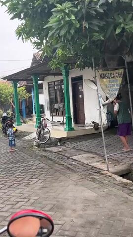 A child in a patterned shirt and blue shorts stands on a brick walkway holding a yellow object. A woman wearing a hijab and a blue patterned dress walks from left to right across the walkway. A motorcycle is parked behind the child. A bicycle is leaning against a white building with green pillars. A cat sits on the steps of the building. A satellite dish is mounted on the side of the building. A broom leans against the building. The walkway is made of red and grey bricks. The building appears to be a single-story structure with a metal roof. Trees are visible behind the building. The scene takes place outdoors.