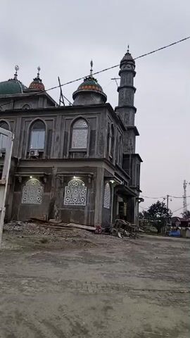 The video pans across the exterior of a mosque that appears to be under construction or recently completed. The building is constructed of gray concrete. It has multiple domes, one large green dome and several smaller, colorful domes. A tall minaret stands to the right of the main building. The windows are arched and some have decorative screens. There is visible construction debris and materials scattered in front of the mosque. The ground in front of the building is unpaved, appearing to be dirt and gravel. The sky is overcast, suggesting it is daytime, possibly early morning or late afternoon. There are no people visible in the immediate vicinity of the mosque. Power lines and a utility pole are visible in the background, along with some trees and what appears to be other buildings in the distance.