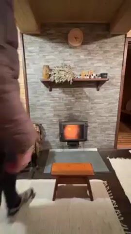 Here's a description of the video: 

The video shows a person in a plaid puffer jacket, dark leggings, and sneakers, and a blue beanie. The person sits on a small, wooden stool in front of a stone-faced fireplace with a burning wood stove. The person reaches to the side and retrieves logs from a firewood rack to the left of the stove.  The environment is a cabin-style room with wood paneling and rugs. The fireplace has a mantle with decorative items. There's a clock on the wall above the mantle. A doorway is visible to the right of the fireplace.