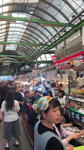 The video shows a bustling indoor market in South Korea. The camera moves slowly through a narrow aisle filled with people, some walking and some sitting at counters eating. There are numerous vendors selling food and other goods from stalls on either side of the aisle. The ceiling of the market has a curved, ribbed design and is partially translucent, allowing natural light to illuminate the scene. There is signage in Korean above the stalls.  Many people appear to be locals, while others look like tourists. Some are casually dressed, others are wearing hats or caps. The general atmosphere is busy and vibrant, typical of a crowded market.