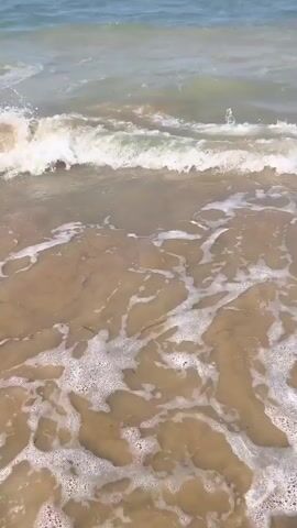 The video shows a close-up, low-angle view of gentle waves washing onto a sandy beach. The water is a murky brownish color, and it is foamy at the edge as it recedes. In the distance, the turquoise ocean stretches out under a bright sky, with some buoys visible on the horizon. The scene is peaceful and natural, with no people or animals clearly visible in the frame. The camera appears to be handheld, with slight movements that track the incoming waves. The overall impression is one of a calm, sunny day at the beach.