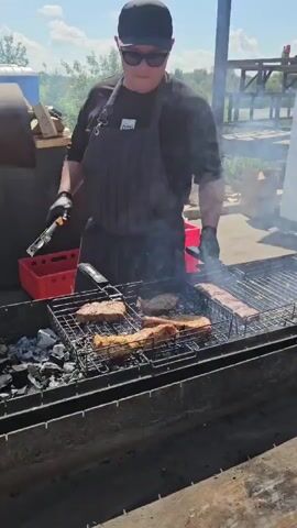 A person wearing a black cap, black sunglasses, a black t-shirt, and a black apron with tongs in their gloved hand is grilling meat over charcoal. The meat is on a grill in front of them. The grill is very long and appears to be a custom-built barbecue pit. There are several pieces of meat on the grill, and smoke is rising from them. The person is using the tongs to turn one of the pieces of meat. In the background, another person wearing a black apron is standing and holding something. There is a red bin to the left of the grill, and a white refrigerator or storage unit behind the people. The scene appears to be outdoors, with trees and a body of water visible in the distance. The ground is concrete or dirt.