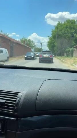 The video is shot from the driver's seat of a car moving down a narrow street. Three cars are visible ahead, in the same direction. The first car is a black Volkswagen Golf, followed by a gray Dodge Challenger with black racing stripes. The third car is a small gray hatchback. The road is bordered by brick buildings on one side and a green tree line with utility poles on the other. The sky is blue with some fluffy white clouds.  The interior of the filming car is visible, focusing on the black dashboard with a textured surface and air vents. The car filming starts moving forward.