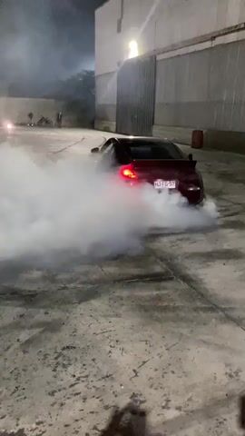 A dark red car is performing a burnout, creating a large cloud of white smoke. The car is in a concrete lot, possibly an industrial area, with a large building in the background. There are tire marks visible on the ground, indicating previous activity. The car's brake lights are illuminated red. The time is nighttime.