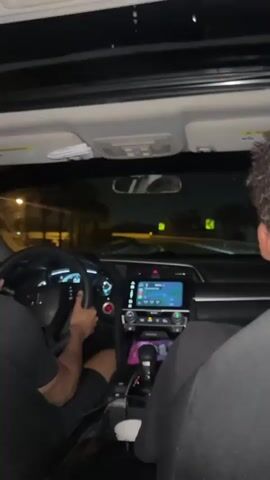 The video shows the interior of a car at night. The driver, wearing a black shirt and shorts, has dark skin and is holding the steering wheel. A passenger is visible in the backseat, wearing a black shirt. The dashboard of the car is illuminated, and a navigation system is displayed on the screen. Street lights are visible through the windshield. The car appears to be moving on a road. The car has a sunroof.