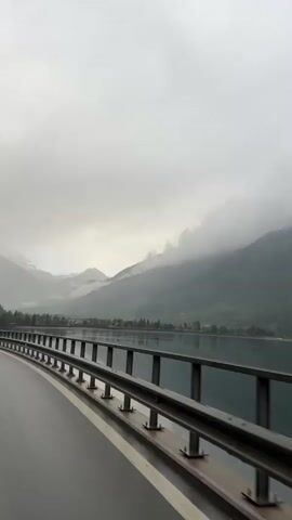 The video, captured from a moving vehicle, showcases a picturesque mountainous landscape in Le Prese, Switzerland, on an overcast day. The foreground features a wet road with a metallic guardrail running along its left side, indicating recent or ongoing rain. Beyond the guardrail, a calm body of water, likely a lake or reservoir, stretches towards the distant mountains.

The surrounding mountains are prominent, with their lower slopes covered in lush green trees. Higher up, the peaks are partially obscured by dense, low-hanging clouds and fog, giving the scene a mysterious and atmospheric quality. Some snow is visible on the highest reaches of the mountains, hinting at their elevation. Scattered small buildings and signs of human settlement can be seen along the shoreline and at the base of the mountains, nestled among the greenery. The overall impression is one of serene, rugged natural beauty under a muted, cloudy sky. No specific people or visible text are present in the frame.