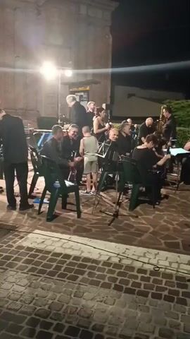 A group of musicians is performing outdoors at night. The musicians, mostly dressed in black, are seated on chairs in front of music stands. A young child stands in the center of the group. One woman in a black dress sings into a microphone. Another woman in a black outfit dances to the music. The background shows a brick building and some greenery. The ground is paved with cobblestones.
