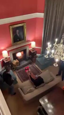 From a high vantage point, the video shows a room with red walls, a fireplace with a fire burning, furniture, and people. A woman in patterned pants and red fuzzy slippers lies on a light-colored sofa in front of the fireplace. A man stands to the left of the sofa, looking at his phone. A portrait hangs above the fireplace. A chandelier hangs from the ceiling. Two chairs, two side tables with lamps, a rug, and a wooden table are also in the room. The camera zooms in closer to the fireplace. Someone calls out "Jerry?"