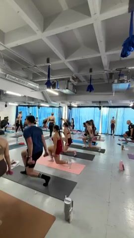 A fitness class is in session in a large, open studio. Approximately 15 people are spread out on yoga mats performing a lunge exercise, some holding small dumbbells. The studio has a high ceiling with exposed ductwork and beams, and blue silk fabrics hang decoratively. A brick wall runs along one side of the room, while the opposite side features a blue curtain. The floor is light gray, and the overall lighting is bright. An instructor stands at the back of the room overseeing the class.