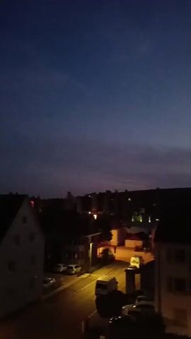 The video pans across a dark suburban landscape at night, under a deep blue sky. Various buildings, mostly in shades of white, red, and dark tones, are visible with some street lighting and car headlights illuminating a street. The scene is slightly out of focus. As the view pans, distant lights on the horizon become visible, suggesting the edge of the town or city. No people are visible in the frame.