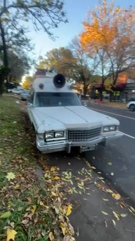The video shows a white, retro-styled "Ghostbusters" themed car parked on the side of a street. The car is a long, boxy model, possibly an old hearse or ambulance that has been customized. It features the "Ghostbusters" logo and lettering prominently displayed on its side, along with black and yellow hazard stripes. The car is equipped with various props and accessories mimicking the equipment from the Ghostbusters movies, including a siren, a large speaker, and other unidentified devices mounted on the roof. The background shows a typical suburban street scene with trees, a sidewalk covered in fallen leaves, and other vehicles parked nearby. The sky is clear and blue, suggesting a sunny day.