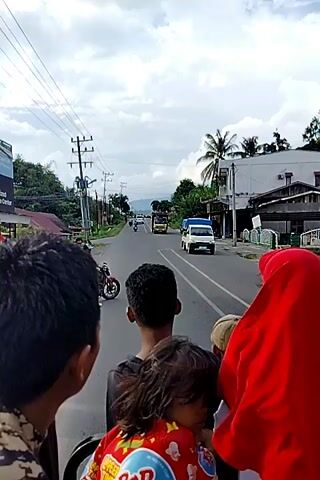 This video shows a rear-view perspective from a vehicle, likely a motorcycle or truck bed, with four individuals visible. The individuals are positioned from foreground to background. In the immediate foreground on the left, the back of a person's head and shoulders are visible. Next to them, slightly more centered and in the mid-ground, is the back of a young boy's head and shoulders. To his right, and partially obscured, is a person wearing a light-colored baseball cap. In the far right foreground, a person wearing a red headscarf and white clothing is looking to the right. Between the boy and the person in the red headscarf, a young child is visible, wearing a red shirt with colorful graphics. The vehicle is traveling on a paved road that extends into the distance, lined with trees, utility poles, and buildings on either side. Several other vehicles, including trucks and motorcycles, are also on the road ahead. The sky is overcast with clouds. The overall impression is a group of people traveling on a road in a semi-urban or rural area.