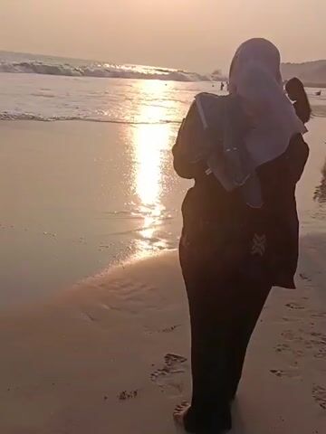 A video shows a person wearing a hijab and dark clothing standing on a sandy beach. The person is facing away from the camera, towards the ocean, where waves are breaking. The sun is setting, casting a warm, golden light across the scene. In the background, another person and a child are visible near the water. The video is approximately 4 seconds long.