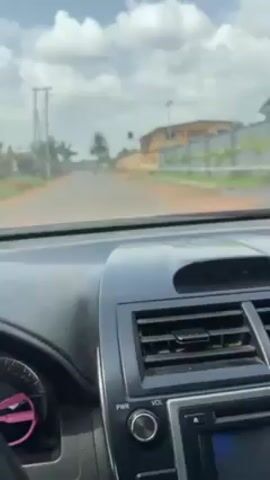The video shows a driver's point of view from inside a car driving down a street. The car has a black dashboard with air vents and a radio. The steering wheel is also black. A hand is visible on the steering wheel. The street is paved and there are buildings and trees on either side. There are also power lines visible. The sky is cloudy with some patches of blue. A motorcycle appears to be further down the road.