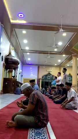 The video shows people praying inside a mosque. The mosque has a red carpet, white and brown patterned ceiling, and decorative architectural features. Many men, mostly wearing casual clothing, are praying on the carpet. An ornate, dark wood structure sits near the left side of the frame. Some men are kneeling with their foreheads on the carpet, while others are sitting and praying. Several ceiling fans hang from the ceiling. A few men stand near the back, observing.