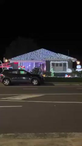 A stationary, eye-level shot captures a house lavishly decorated with Christmas lights at night. The house, a one-story structure with a light-colored facade, is entirely covered in lights; the roof is draped with white lights, and the yard is adorned with colorful holiday decorations, including a "Ho Ho Ho" sign. A dark gray SUV is parked on the street in front of the house, partially obscuring the view of the holiday display. The street itself is well-lit, and the background is dark with a few trees visible.